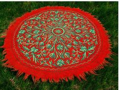 a red and green round rug sitting in the middle of some grass with an intricate design on it