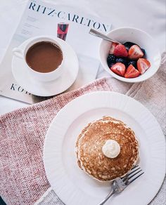 a white plate topped with pancakes and fruit next to a cup of coffee on top of a table