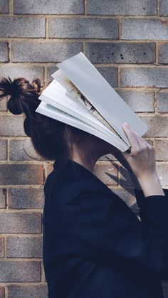 a woman with her hair in a bun holding books up to her face while standing against a brick wall