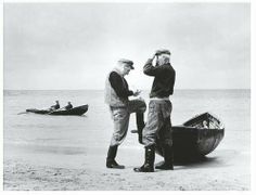 two men standing next to each other near boats on the beach with one man looking at his cell phone