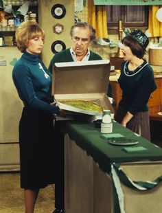 two women and an older man are looking at a pizza in a box on a table