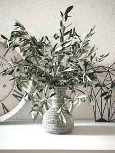 a vase filled with green leaves sitting on top of a white shelf next to two clocks