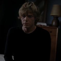 a young man with blonde hair sitting on a bed in a dark room looking at the camera