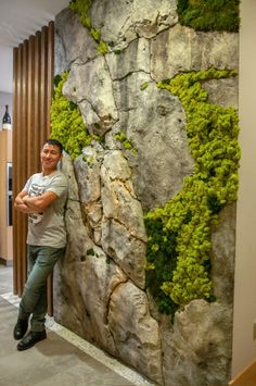 a man standing next to a wall covered in green mossy plants and rocks with his arms crossed