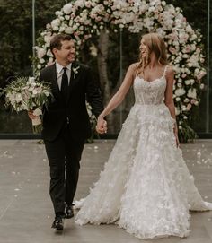 a bride and groom holding hands while walking through an archway decorated with flowers in front of them