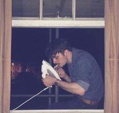 a man sitting on a window sill holding a white object in his hand and looking out the window