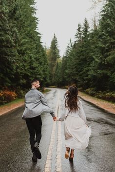 a man and woman holding hands while walking down the road in the rain with trees behind them