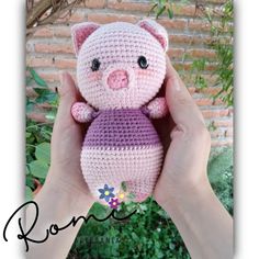 a crocheted pink and purple pig is held in front of a brick wall