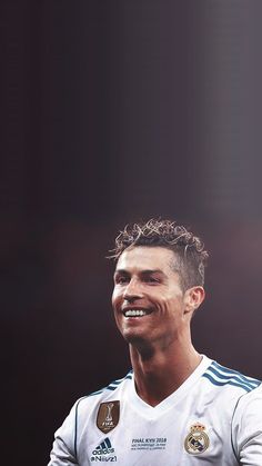 a man with curly hair smiles while wearing a white shirt and blue shorts, standing in front of a black background