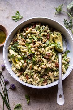 a white bowl filled with pasta salad next to two spoons and some green herbs