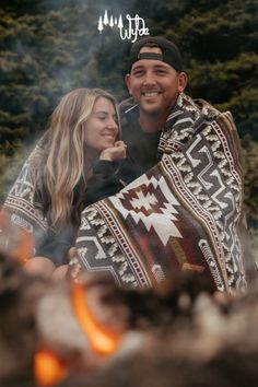 a man and woman sitting next to each other near a campfire in the woods