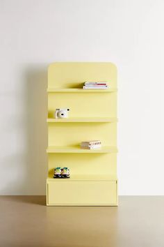 a yellow book shelf with three shelves and two toy cars on top of it, in front of a white wall