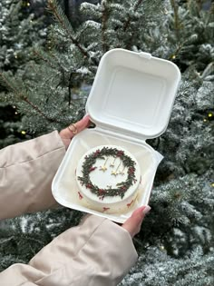 a person holding a cake in a box with wreath decorations on it while standing next to a christmas tree