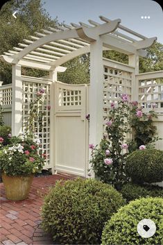 a white arbor with pink flowers in it and some bushes around it on a brick walkway