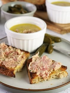 two pieces of bread on a plate with some soup and pickles in the background