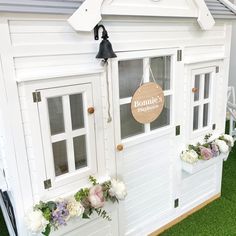 a white doll house with flowers on the front and side windows, sitting in grass