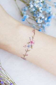 a small flower tattoo on the left wrist and right arm, with blue flowers in the background