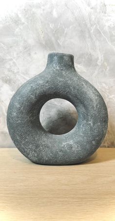 a gray vase sitting on top of a wooden table next to a white wall with a black circle