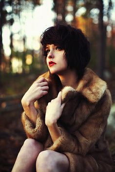 a woman in a fur coat sitting on the ground with her hand under her chin