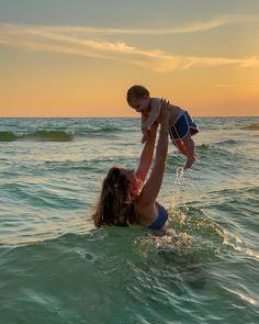 a woman holding a baby in the ocean at sunset with waves crashing on her head