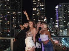 three women posing for the camera at night in front of city lights and skyscrapers