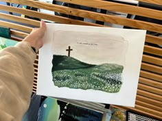 a person holding up a painting with a cross on it in front of a bench