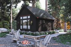 a small black cabin sits in the middle of a wooded area with chairs around an open fire pit