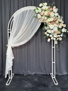 a white wedding arch decorated with flowers and sheer drapes for an outdoor ceremony in front of a gray backdrop