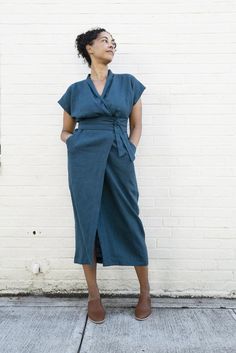 a woman standing in front of a white brick wall wearing a blue dress and brown shoes