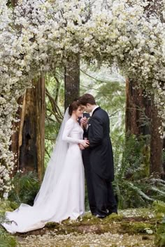 a bride and groom standing in front of an archway with white flowers on the trees