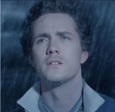 a man with curly hair and blue eyes stares into the distance while standing in the rain