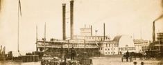 an old photo of people and horses in front of a factory with smokestacks