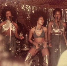 three women singing into microphones while standing in front of a stage with other people