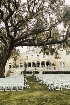 an outdoor ceremony setup with white chairs and trees in front of a large building on the lawn