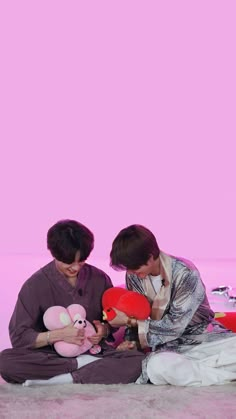 two young men sitting on the ground with stuffed animals in front of pink background, one holding a teddy bear