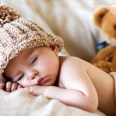 a baby is sleeping on a bed with a teddy bear next to it and wearing a knitted hat