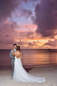 a bride and groom standing on the beach at sunset