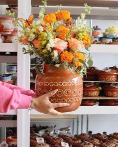 a person holding a vase with flowers on it in front of shelves full of cakes