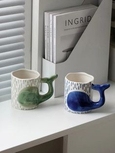 two ceramic mugs sitting on top of a white counter next to a book shelf