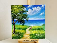 an easel with a painting of a path leading to the ocean and flowers on it