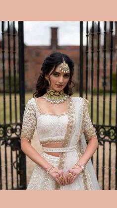 a woman standing in front of a gate wearing a white and gold lehenga