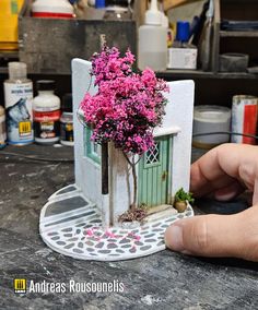 a hand is holding a miniature house with pink flowers in the front and behind it