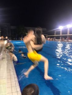 two people are jumping into the pool at night, one is wearing yellow swim trunks