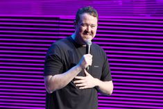 a man standing in front of a purple wall holding a microphone to his chest and smiling at the camera