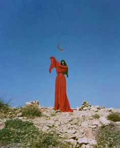 a woman in a long red dress standing on top of a rocky hill with a crescent above her head