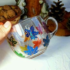 a hand is holding a glass cup with leaves painted on it and next to some pine cones