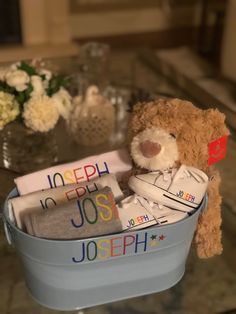 a teddy bear sitting in a bucket filled with personalized baby items on a table