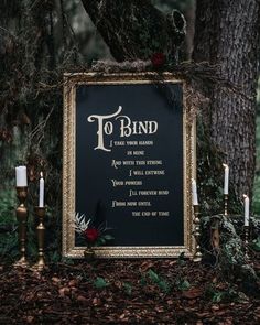 a black and gold framed sign sitting in the middle of a forest next to candles