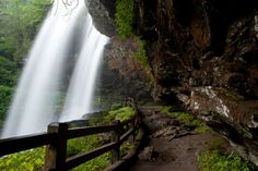 a large waterfall in the middle of a lush green forest next to a wooden fence