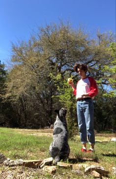 a woman standing next to a black dog on top of a grass covered field with trees